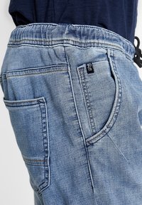 Joggers de mezclilla azul claro con cintura elástica, bolsillos laterales y una pequeña etiqueta negra con un logo en el bolsillo trasero.
