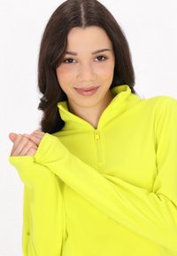 Fluorescerende gul zip-up fleece, blød tekstur, høj krave, lange ærmer med tommelfingerhuller, frontlynlås, båret af en person med hænderne flettet.