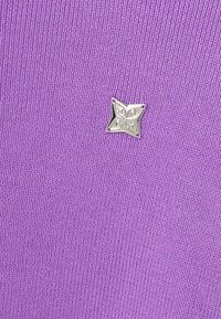 Un tissu tricoté violet présente un logo en forme d'étoile à quatre pointes argentée avec une surface texturée, positionné de manière proéminente sur le vêtement.