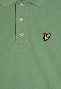 Grønn polo skjorte laget av teksturert bomull med en tre-knapps placket og en gul og svart brodert logo på venstre bryst.
