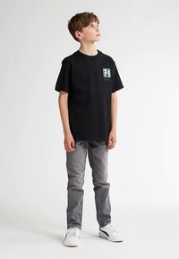 Tricou negru din bumbac cu mâneci scurte, având un mic desen colorat pe piept, asortat cu blugi gri și adidași albi.