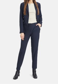 Mörkblå skräddarsydd kostym med en figursydd kavaj och smala byxor, tillsammans med en krämfärgad topp och svarta ankelboots. Slät tygstruktur.