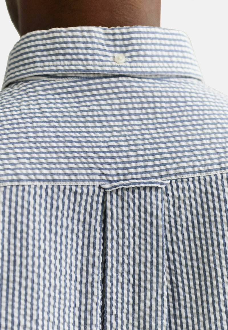 Primer plano de una persona que lleva una camisa con rayas verticales en azul claro y blanco, con el cuello trasero abotonado.