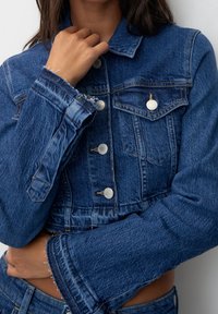 Chaqueta de mezclilla azul recortada que presenta cierres de botones plateados, dos bolsillos en el pecho y bordes de manga deshilachados, destacando un acabado texturizado.