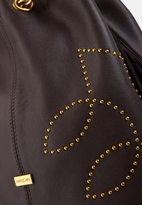 Braune Ledertasche mit goldenen Nieten, die ein Blattmuster bilden. Verfügt über ein goldenes Logo-Schild und eine strukturierte Oberfläche. Zugverschluss und weicher Fall.