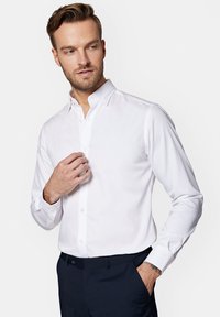 Biała koszula z guzikowym kołnierzykiem, długimi rękawami i zapięciem na przodzie. Gładka faktura materiału, dopasowany krój i subtelny połysk.