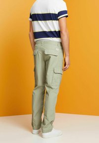 Man in beige cargobroek, marineblauw en wit gestreept shirt met korte mouwen, en witte sneakers, staand tegen een oranje achtergrond.
