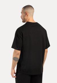 Svart bomullströja med avslappnad passform, korta ärmar och rak nertill. Synlig tatuering på armen, minimalistisk design utan logotyper.