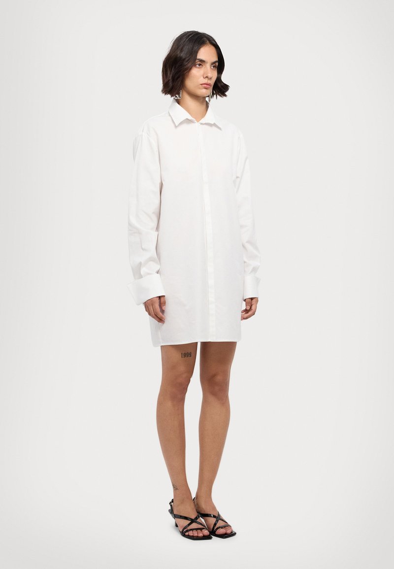 Vit oversized skjortklänning med knappfront, långa ärmar och uppvikta manschetter. Buren över benen, med en tatuering på låret.