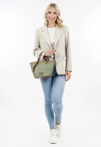 Borsa a tracolla verde intrecciata con dettagli in pelle marrone, manici superiori e un design strutturato. Il modello indossa jeans chiari e un blazer beige.