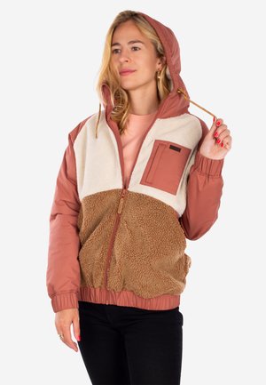 Frau, die die Kordel an einer Kapuzenjacke mit braunen und cremefarbenen Fleeceteilen, rosafarbenen Ärmeln und Brusttasche anpasst, trägt schwarze Hose.