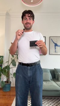 Μαύρο υφασμάτινο πορτοφόλι κρατημένο σε ένα χέρι, φορεμένο με λευκό μπλουζάκι και φαρδιά μπλε τζιν. Το φόντο περιλαμβάνει πράσινο φυτό και καναπέ σε ανοιχτόχρωμους τόνους.