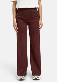 Frau in hochtaillierten, weit geschnittenen bordeauxfarbenen Hosen mit Faltendetail, einem weißen, eingesteckten Hemd und silbernen Turnschuhen, die vor einem weißen Hintergrund steht.