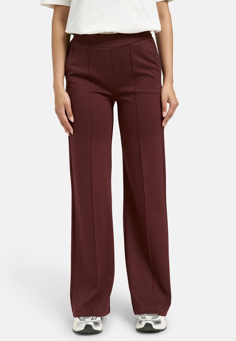 Frau in hochtaillierten, weit geschnittenen bordeauxfarbenen Hosen mit Faltendetail, einem weißen, eingesteckten Hemd und silbernen Turnschuhen, die vor einem weißen Hintergrund steht.