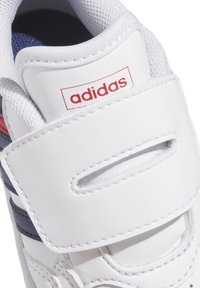 Scarpa sportiva bianca con tomaia in pelle e rete, dotata di accenti rossi e blu, chiusura con strap in Velcro e dettagli cuciti.