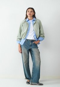 Jasnozielona kurtka bomberka na niebieskiej koszuli w paski, białej koszulce i wyblakłych dżinsach typu wide-leg, w zestawieniu z srebrnymi balerinami.