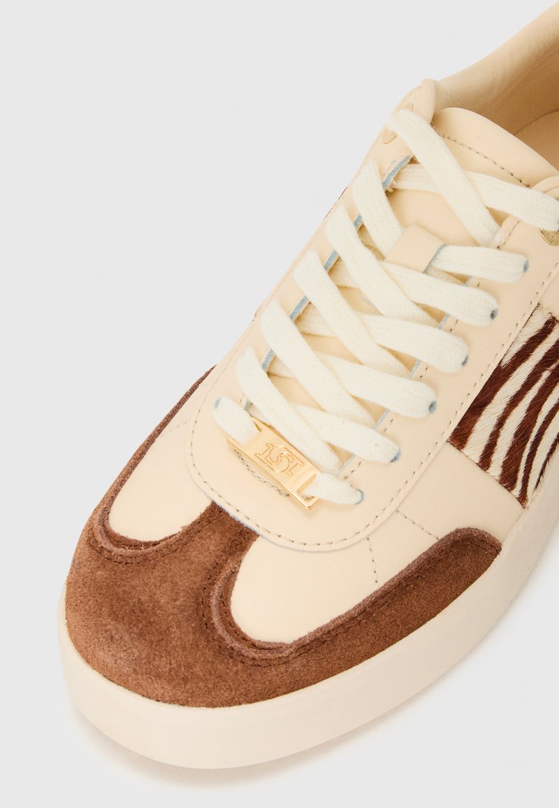 Baskets beiges avec un capuchon en daim marron, lacets crème et détail latéral à rayures zébre sur fond blanc.