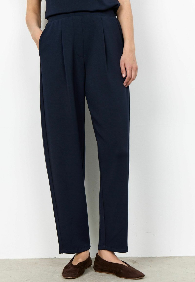 Personne portant un pantalon plissé bleu marine avec les mains dans les poches et des ballerines en daim marron, debout contre un mur blanc.