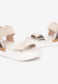 Sandales à glissière beiges et métalliques avec une épaisse semelle blanche, dotées de brides croisées et d'un accent léopard à l'arrière.