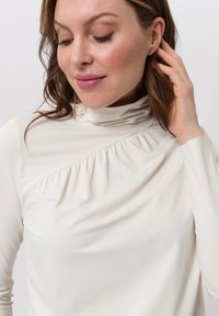 White high-neck top with gathered fabric detail on one side, long sleeves, and a smooth, soft texture.
