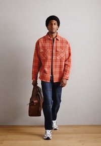 Camisa de franela a cuadros en tonos naranja y rojo, con dos bolsillos en el pecho, combinada con vaqueros azules y zapatillas blancas. Incluida una bolsa de cuero marrón.