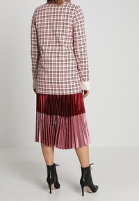 Blazer a scacchi con colletto bianco e polsini a coste; abbinato a una gonna plissettata color block in tonalità rosse e rosa; stivali neri alla caviglia.