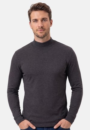 Mand med lyst brunt hår iført mørkegrå langærmet skjorte med høj hals, stående med hænderne i lommerne mod en ensfarvet lys baggrund.