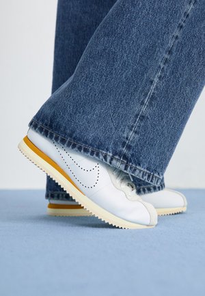 White leather sneakers with yellow accents, round toe, and textured rubber sole, paired with dark blue denim flared pants.