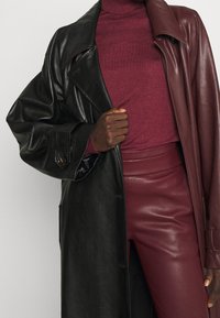 Abrigo de cuero negro con un gran cuello, combinado con un suéter de cuello alto burdeos y pantalones burdeos de talle alto y brillantes.