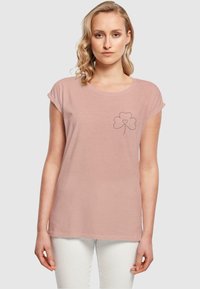 Lyserød T-shirt med korte, optrukne ærmer. Har et sort omrids af en kløver med et hjerte i midten på venstre bryst.