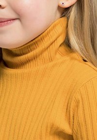 Zbliżenie na osobę noszącą musztardowo-żółty sweter z prążkowanej dzianiny z golfem i małym srebrnym kolczykiem z czerwonym serduszkiem.