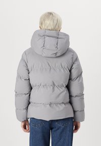 Ljusgrå puffjacka med en tjock huva, quiltat mönster och elastiska ärmar. Plagget har en mjuk textur och bärs med blå jeans.