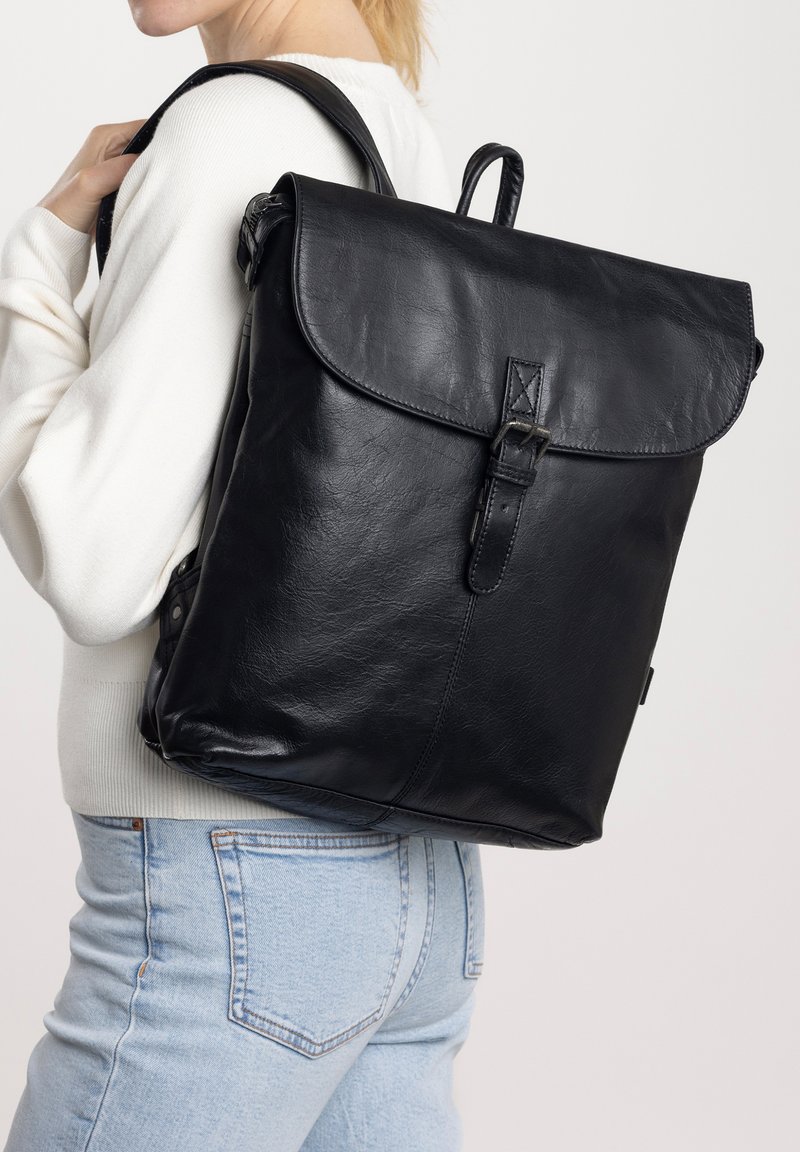 Zaino in pelle nera con chiusura a pattella, dettagli in fibbia e superficie texture. Cinghie per le spalle e maniglia superiore visibili su una persona.