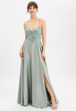 Femme portant une robe en satin vert clair avec un corsage floral orné de perles, des bretelles fines et une fente haute sur la cuisse, debout devant un fond blanc uni.