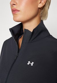 Chaqueta negra con cremallera y cuello alto, con un logo blanco en el lado izquierdo, hecha de un material suave y elástico.