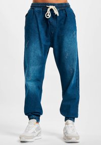 Pantalones jogger de mezclilla azul con una cinturilla elástica, cordón ajustable y tobillos acampanados. Presenta acentos desgastados y bolsillos laterales.