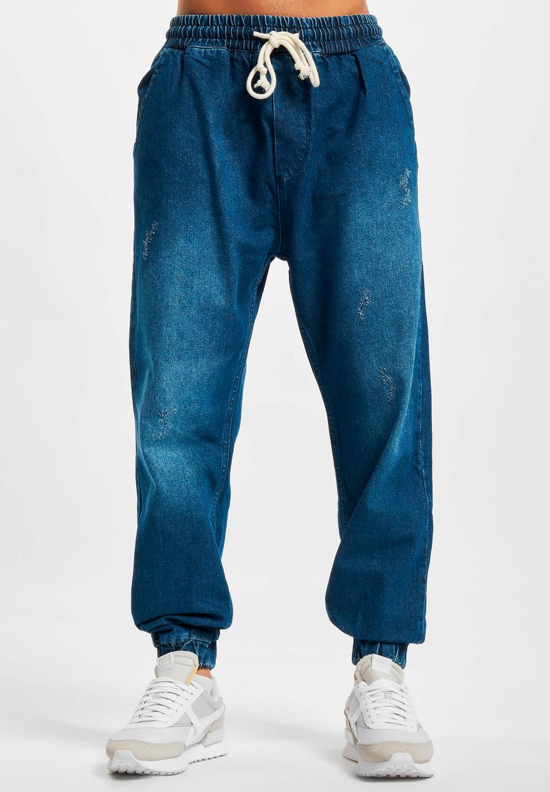 Pantalones jogger de mezclilla azul con una cinturilla elástica, cordón ajustable y tobillos acampanados. Presenta acentos desgastados y bolsillos laterales.
