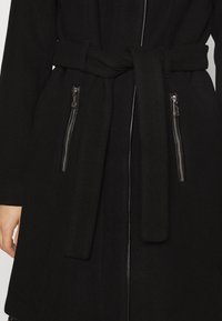 Cappotto di lana nera con vita a cintura, dotato di chiusura lampo frontale e due tasche laterali con zip. Texture liscia con vestibilità sartoriale.