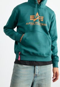 Felpa color teal in materiale morbido, con tasca a marsupio. Presenta un testo logo arancione e dettagli, polsini a costine e un'etichetta rossa sull'orlo.