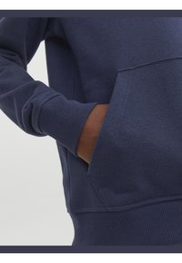Tmavě modrý svetr s ribbed manžetami a lemem, měkkou texturou, klokaní kapsou a prošívanými detaily na švech.
