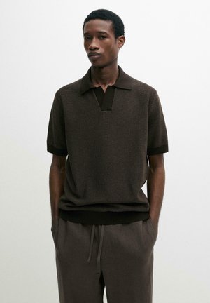 Mann trägt dunkles, strukturiertes kurzärmeliges Polohemd und lockere dunkle Hose mit den Händen in den Taschen, stehend vor einem einfarbigen hellen Hintergrund.