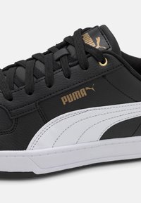 Black leather sneaker with white stripe, gold branding, and black laces. Features a rubber sole and textured surface.