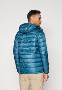 Teal puffer jakna s kapuljačom, koja ima tkanu šaru, sjajnu teksturu i logotip na leđima, uparena sa bež hlačama.