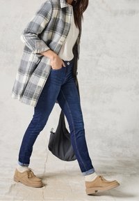 Grå och vit rutig kappa, vit skjorta, mörkblå jeans och bruna skor. Modell som går, med en svart väska hängande över axeln.