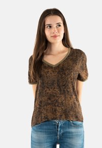Camiseta marrón de cuello en V con un patrón texturizado y ribete verde oliva. Mangas cortas y un ajuste relajado; combinada con jeans azules.