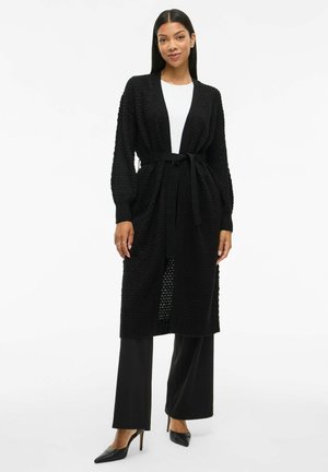 Cardigan noir en tricot avec un tissage texturé, devant ouvert et ceinture assortie. Associé à un pantalon large noir et des escarpins pointus.