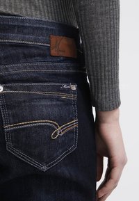 Jeans in denim scuro con tasca posteriore curva, cuciture dorate e etichetta in pelle marrone in vita, abbinati a una maglia grigia a coste.