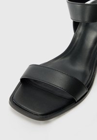Sorte lær sandaler med hæl som har to brede stropper, åpen tå og firkantet form. Glatt tekstur med minimale dekorasjoner.