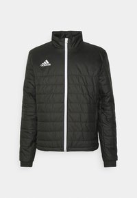 Sort polstret jakke med høj krave, hvid lynlås og hvid Adidas-logo på brystet. Har quiltet tekstur og ribbede manchetter.