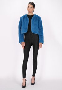 Giacca cropped in pelliccia sintetica di un blu vivace, caratterizzata da un collo rotondo e maniche lunghe. Indossata sopra una maglia nera e abbinata a leggings neri lucidi.